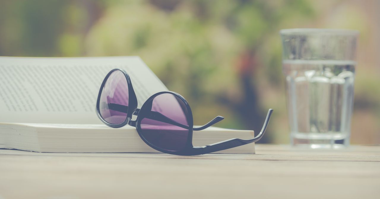 Sunglasses resting on an open book with a glass of water in an outdoor setting. Perfect for leisure and relaxation themes.
